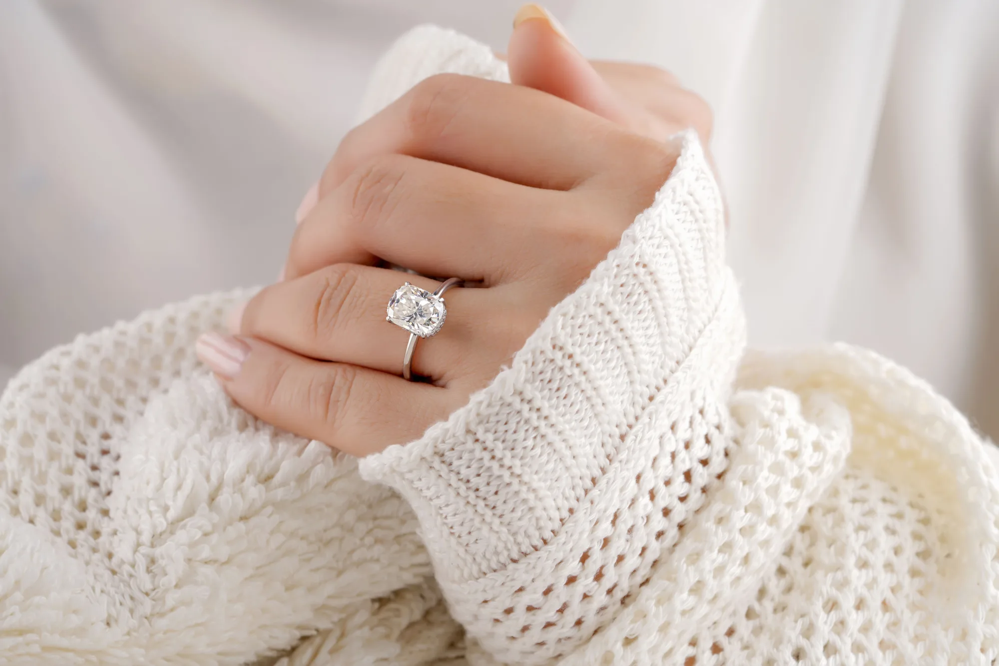 A hand wearing an elongated cushion-cut diamond solitaire ring, against a cream knit sweater